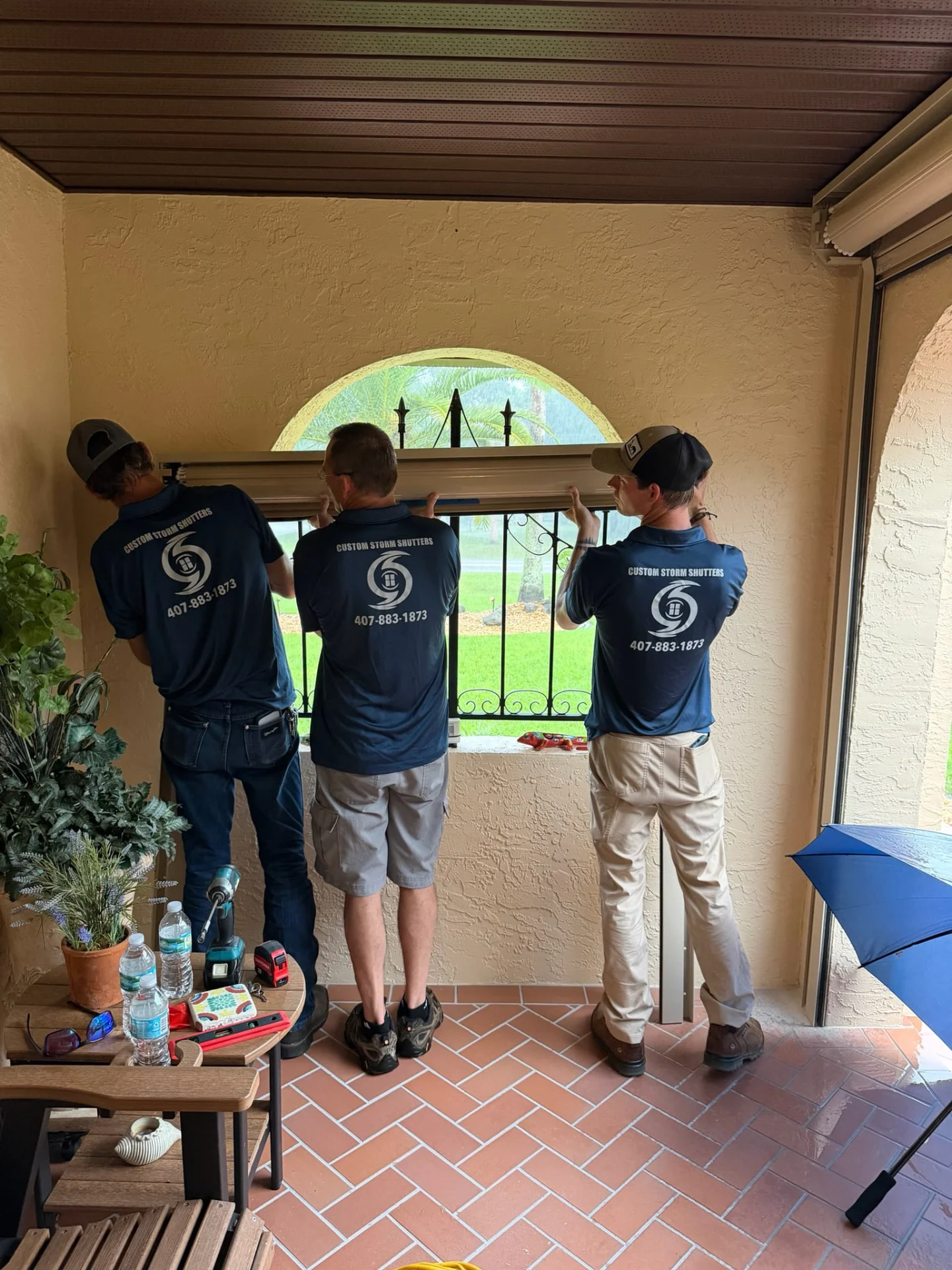 Three Custom Storm Protection installers in branded polo shirts fitting a tan roll-down shutter component over an arched window from inside a porch