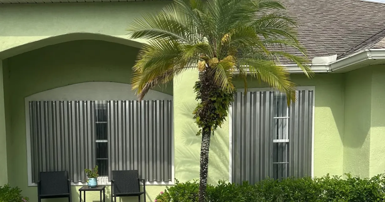 Corrugated storm panels installed over the front windows of a green stucco home